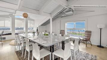 A spacious dining area with a long table, white chairs, and a view of the beach.