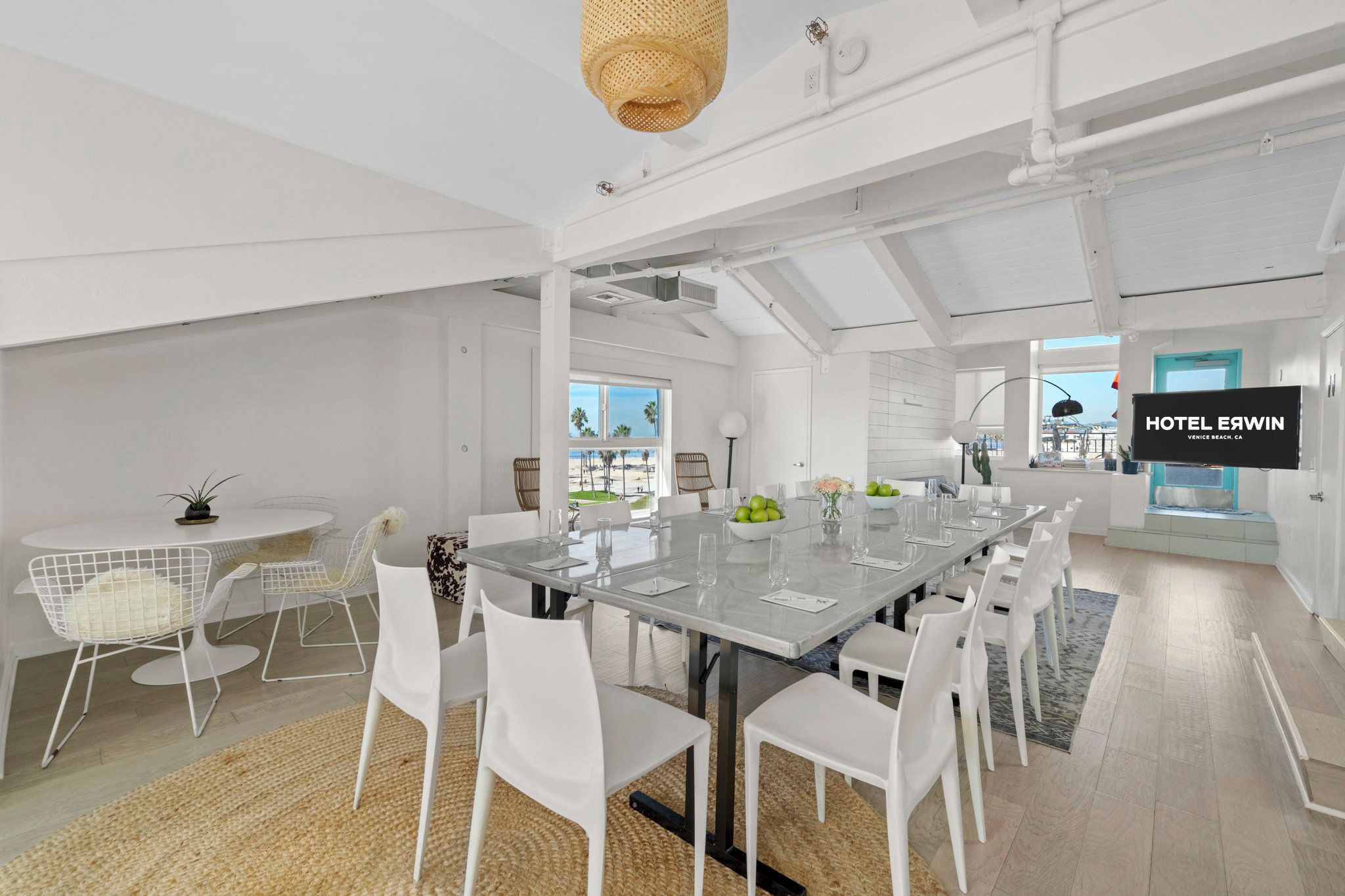 A modern hotel room with a large dining table, white chairs, a TV, and a view of the beach.