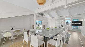 A modern hotel room with a large dining table, white chairs, a TV, and a view of the beach.