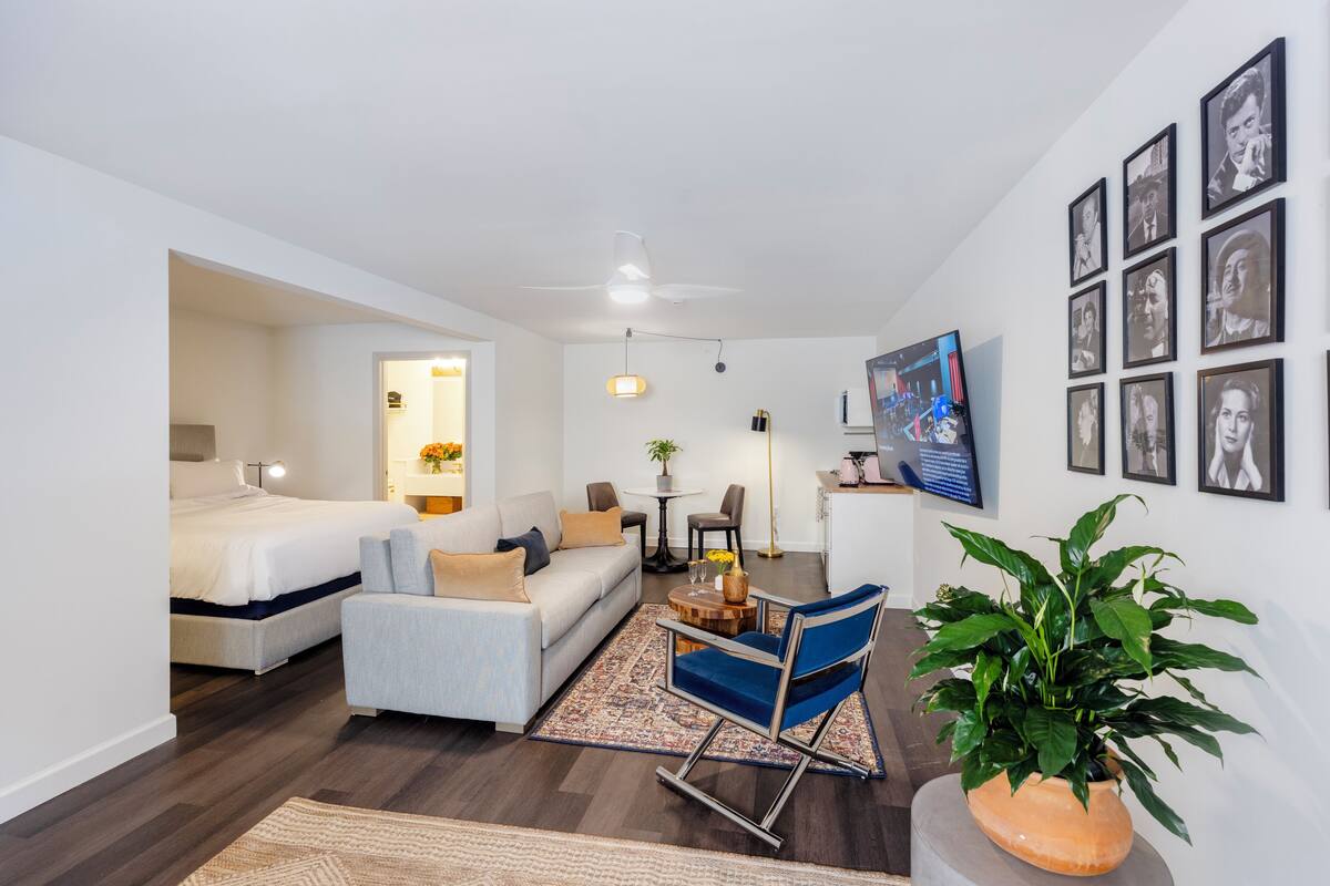 A modern hotel room with a bed, a sofa, a dining area, and a living area. There are framed pictures on the wall and a plant in the corner.