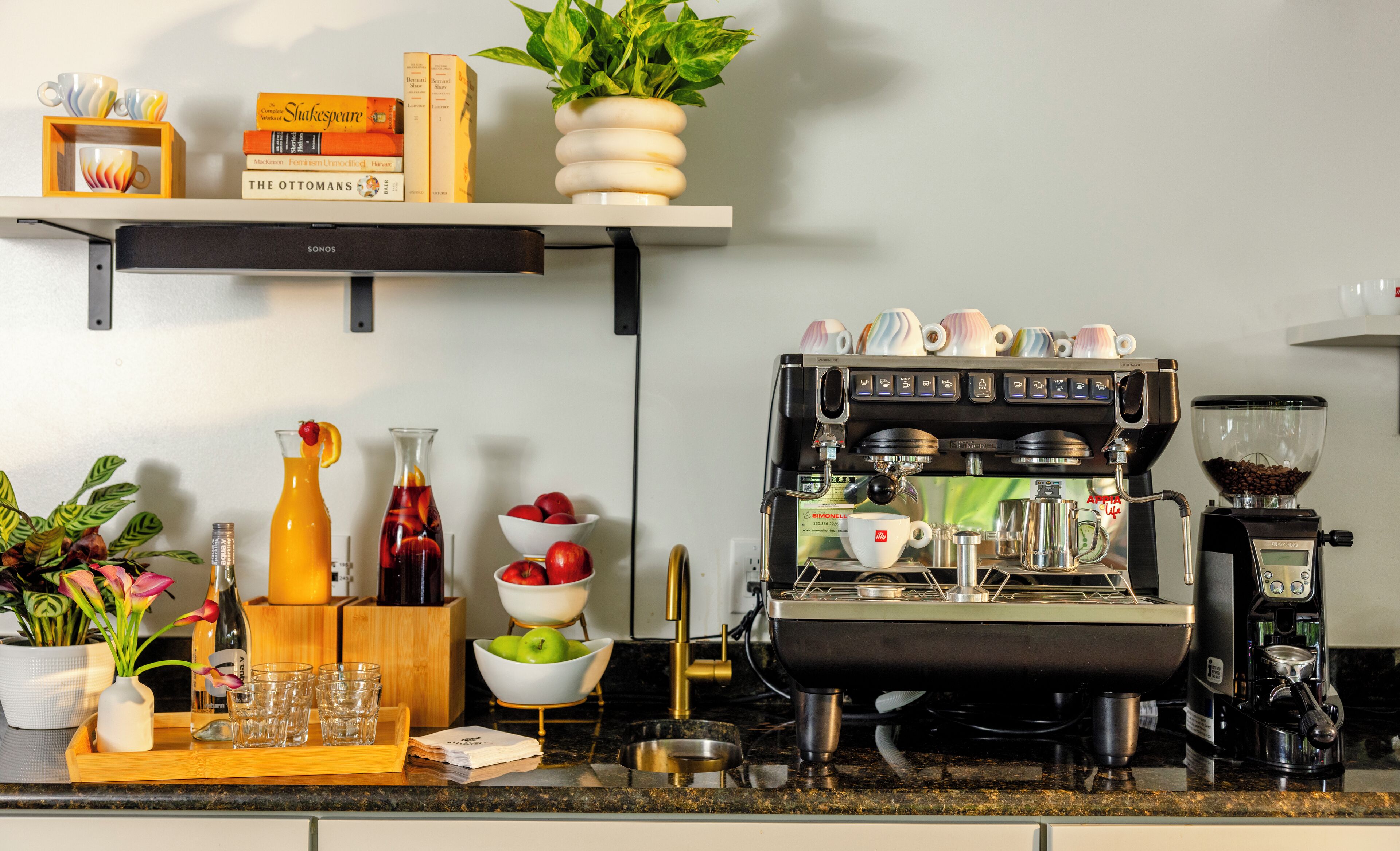 Una encimera de cocina con una máquina de espresso, varios utensilios de cocina y una estantería con libros y una planta.