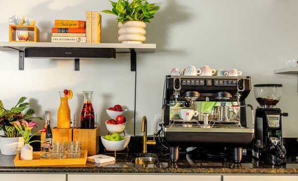 Una encimera de cocina con una máquina de espresso, varios utensilios de cocina y una estantería con libros y una planta.