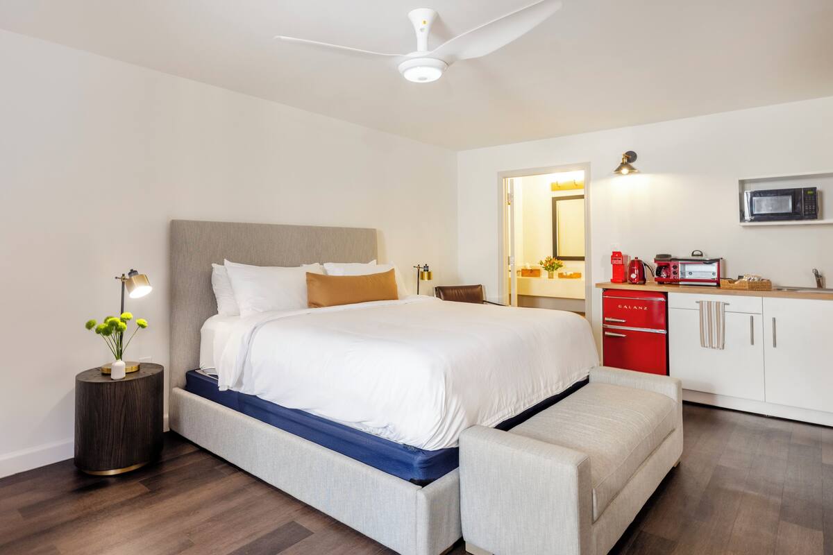 A modern hotel room with a large bed, a small kitchenette, and a ceiling fan.