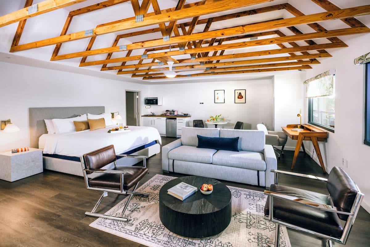 A modern hotel room with a large bed, a sofa, a dining table, and a kitchen area.