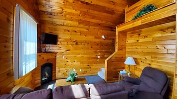 Interior acogedor de una cabaña con paredes de madera, chimenea, sofá, televisor y una pequeña escalera.