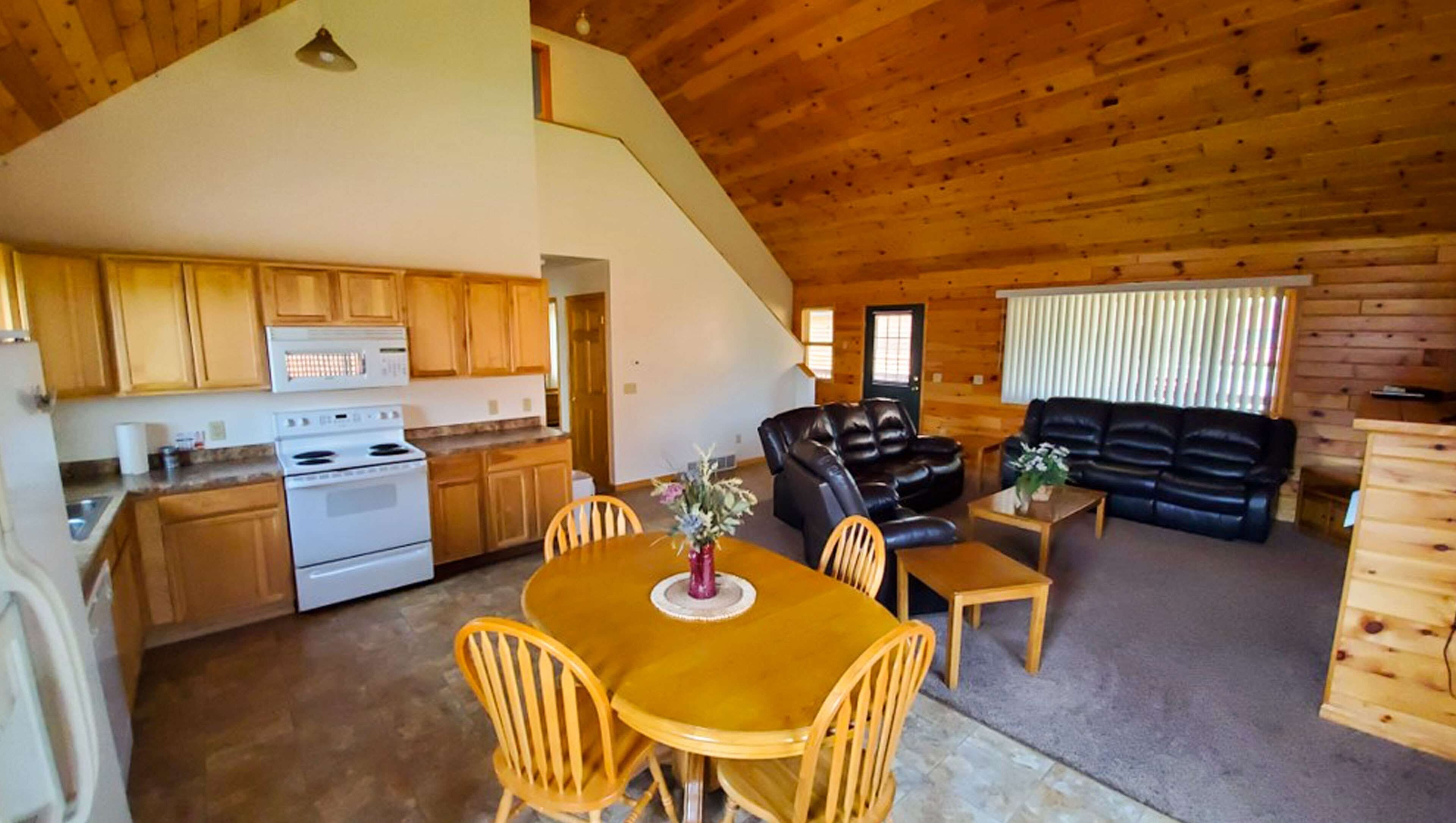 A spacious living area with a wooden ceiling, a dining table with chairs, a sofa, and a kitchen with wooden cabinets and a stove.