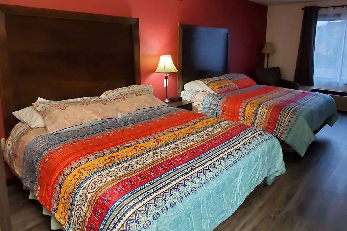 A hotel room with two beds, a wooden headboard, a nightstand with a lamp, a desk, and a window with curtains.