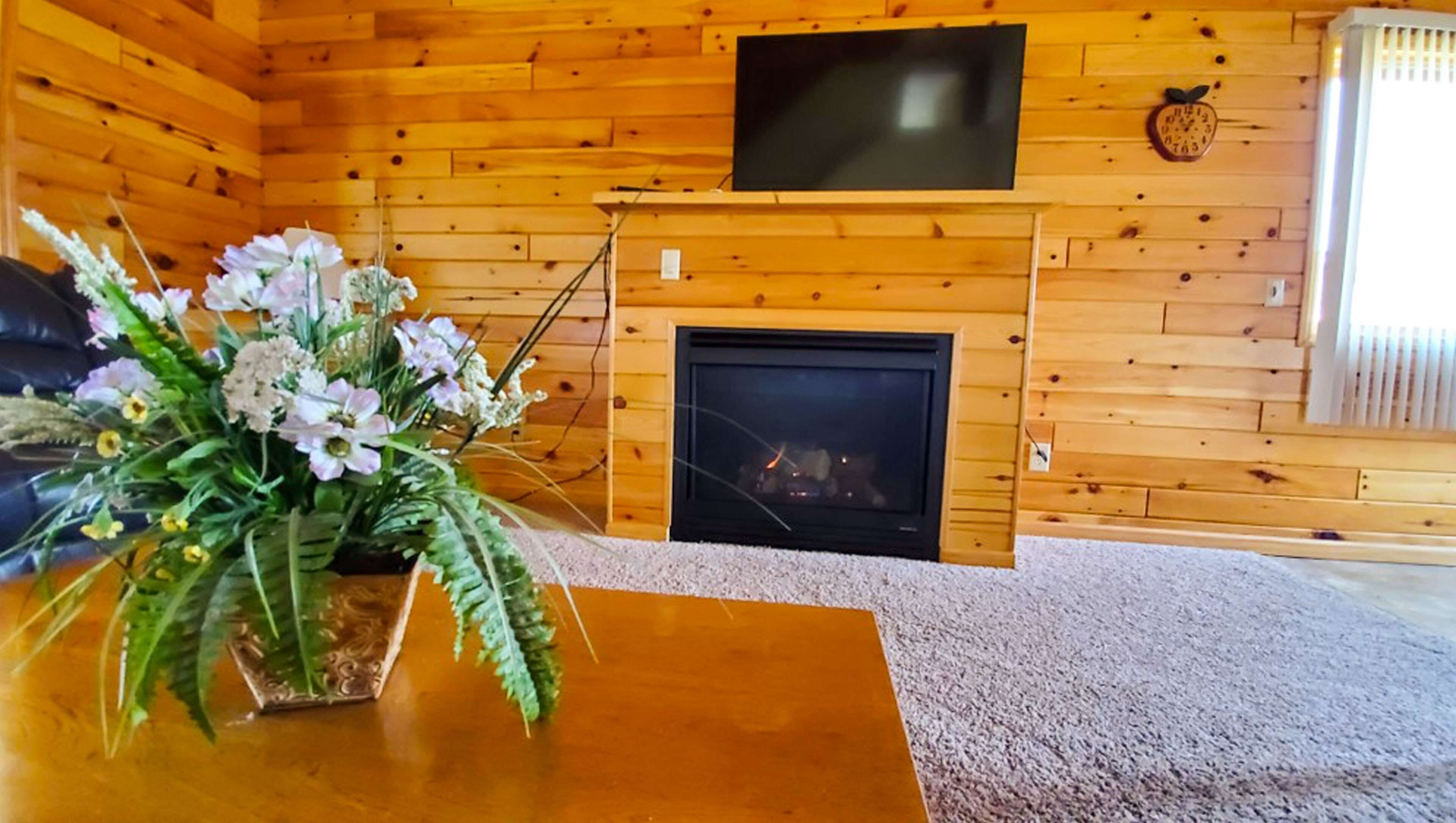 Une pièce chaleureuse avec une cheminée, un téléviseur à écran plat fixé au-dessus, et un arrangement floral sur une table en bois.