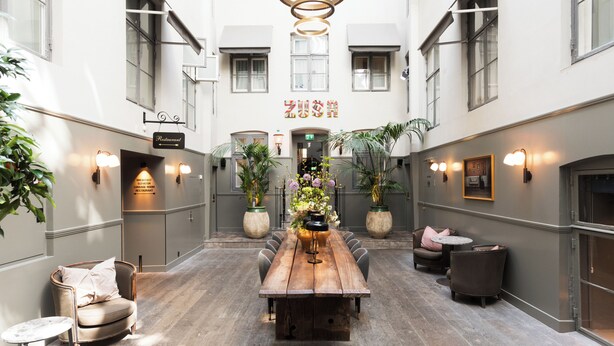 A spacious indoor area with a long wooden table, several chairs, potted plants, and wall-mounted lighting fixtures. - Hotel SKT. Annæ (Copenhagen)