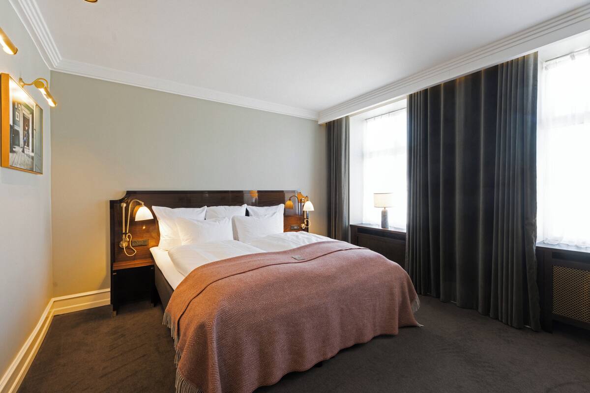 A hotel room with a large bed, a nightstand with a lamp, a window with curtains, and a wall-mounted picture.