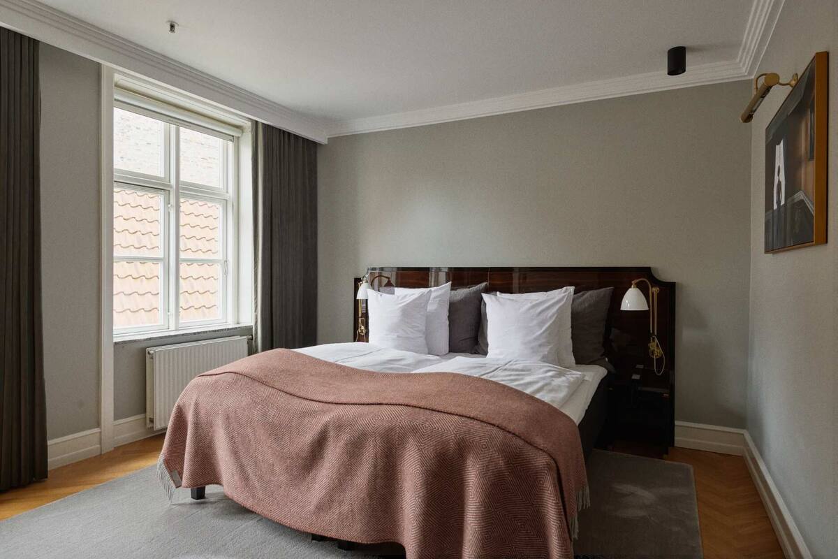 A neatly arranged bedroom with a large bed, a window with curtains, a radiator, and a framed picture on the wall.