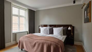 Een keurig opgemaakte slaapkamer met een groot bed, een raam met gordijnen, een radiator en een lijst met een afbeelding aan de muur.