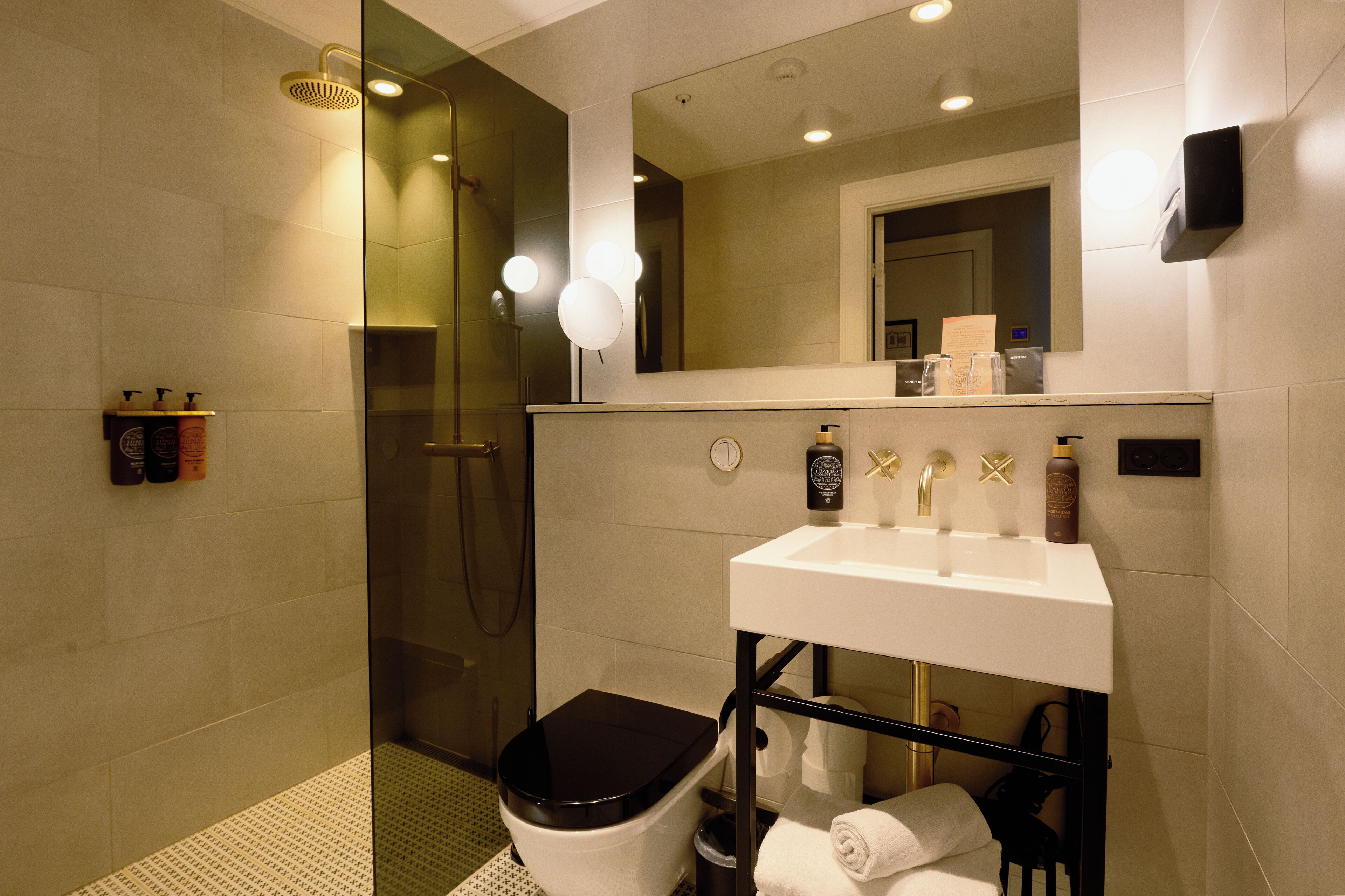Un baño moderno con ducha envuelta en vidrio, un lavamanos blanco con detalles dorados y un inodoro negro.