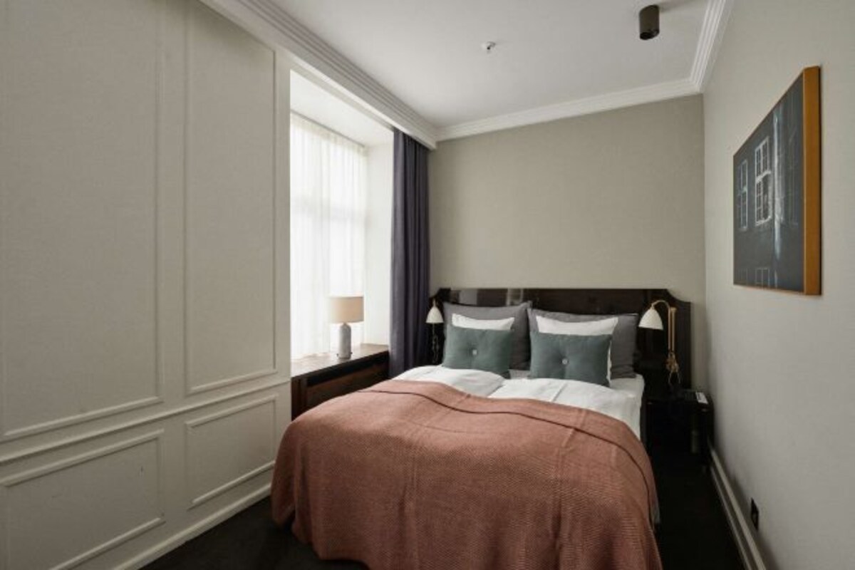 A hotel room with a large bed, a nightstand, a window with curtains, and a painting on the wall.