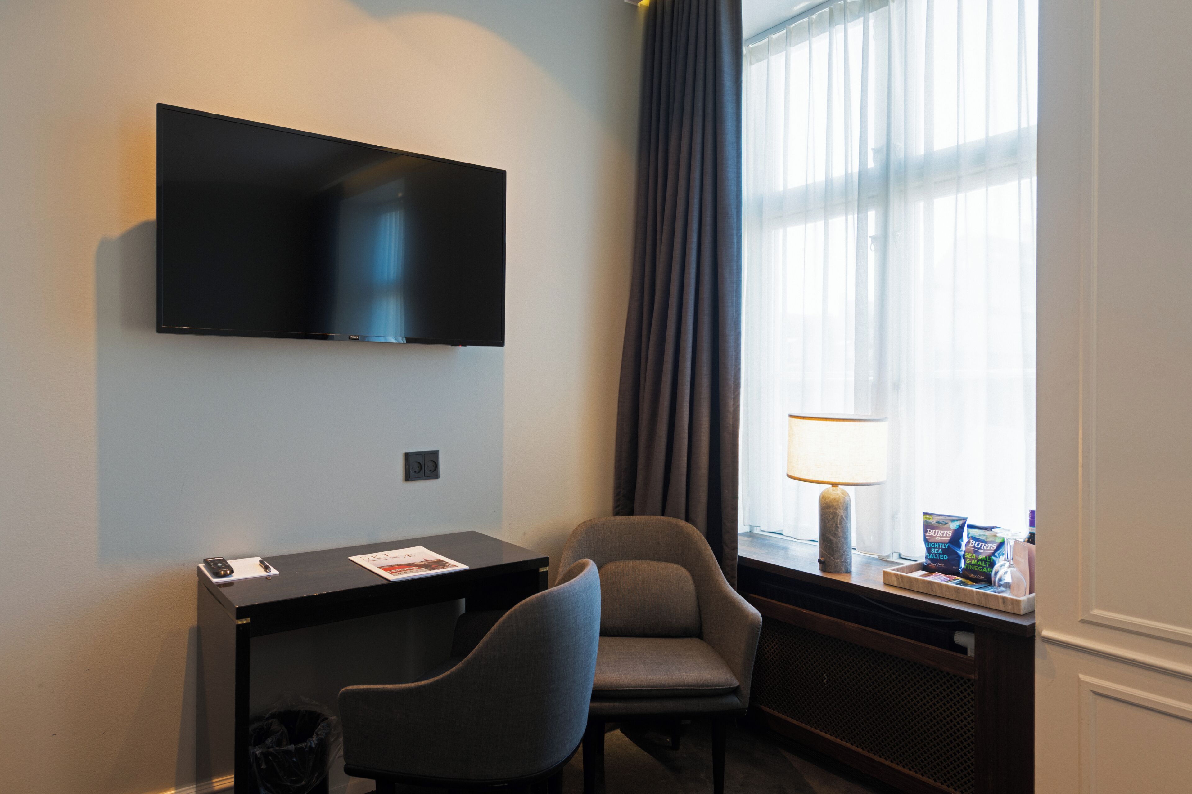 Quarto de hotel com televisão de tela plana na parede, uma mesa com cadeira, janela com cortinas e luminária no parapeito.