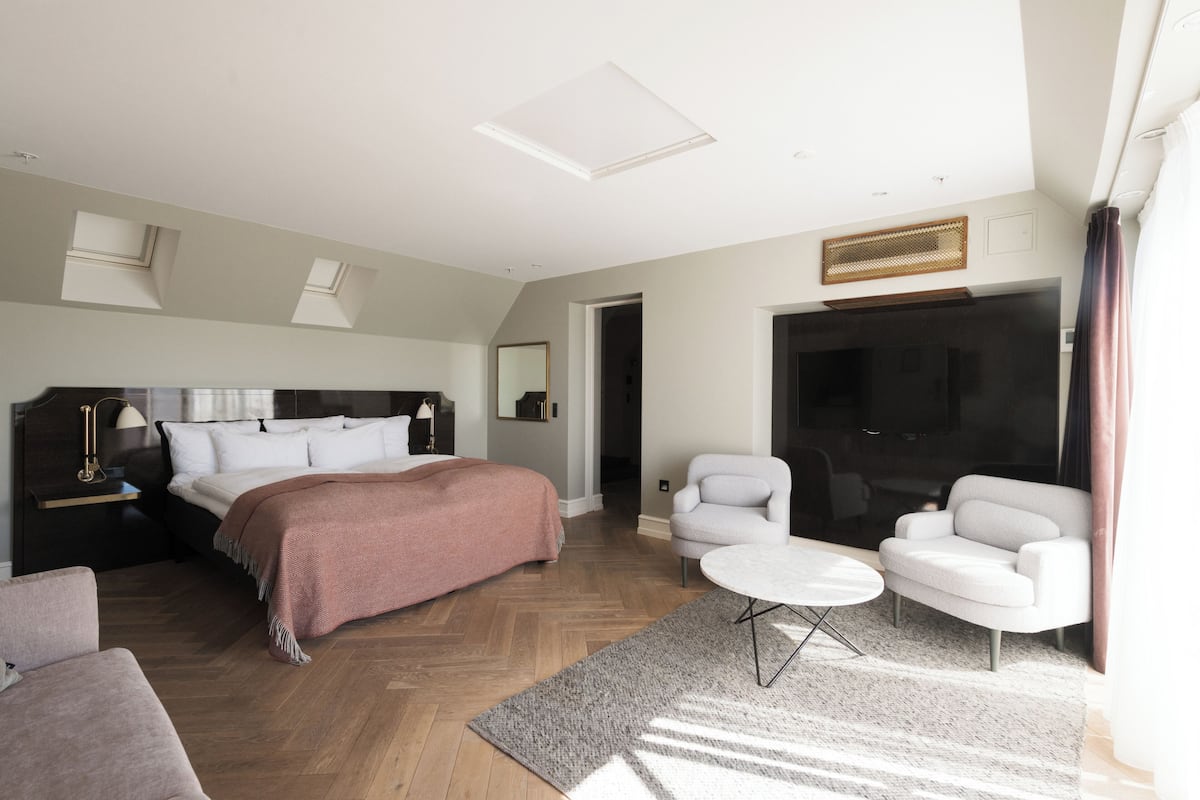 A modern hotel room with a large bed, two white armchairs, a small round table, a TV, and a large window.