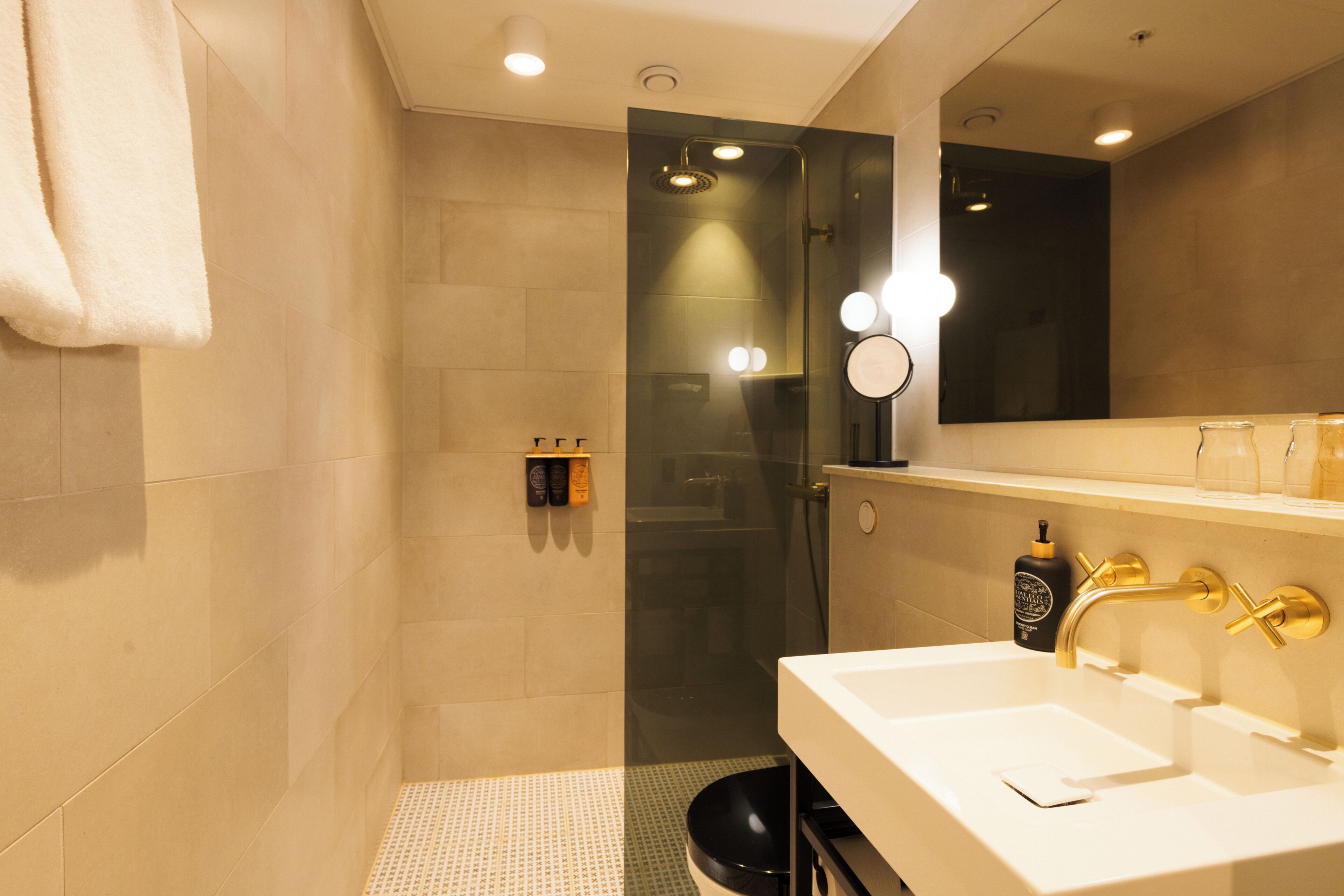 A modern bathroom with a glass shower enclosure, a white sink with gold fixtures, and a mirror with lights.