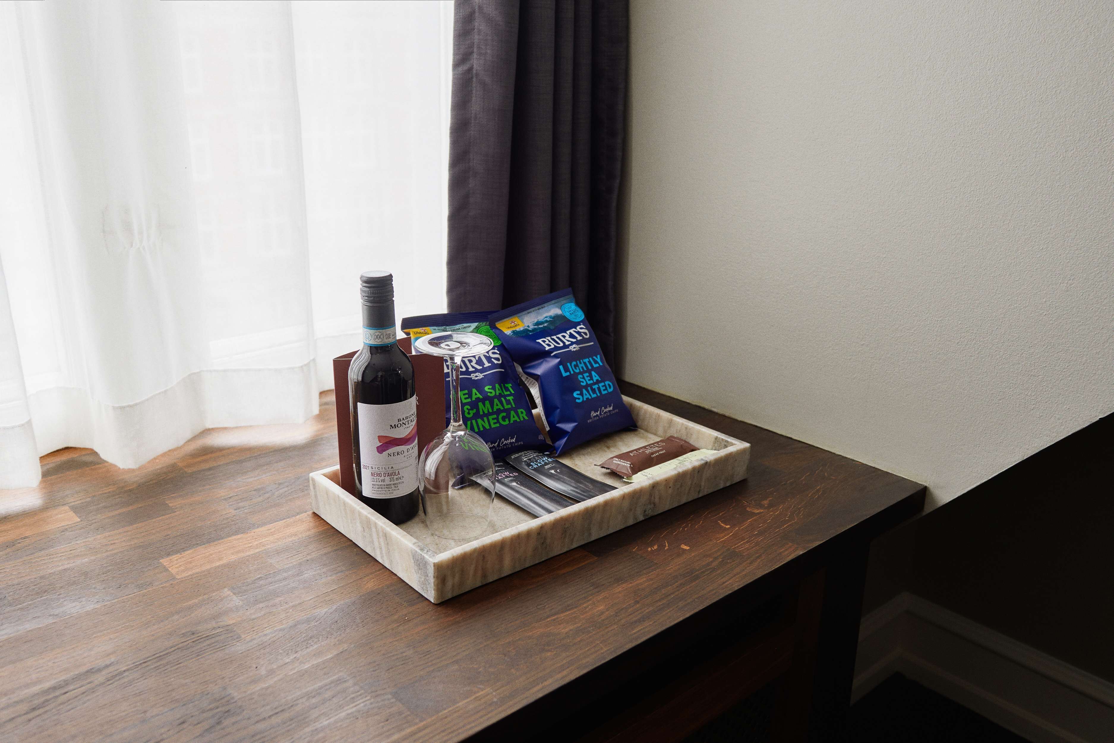 A tray with a bottle of wine, a glass, and bags of sea salt and lightly salted snacks on a wooden table by a window.