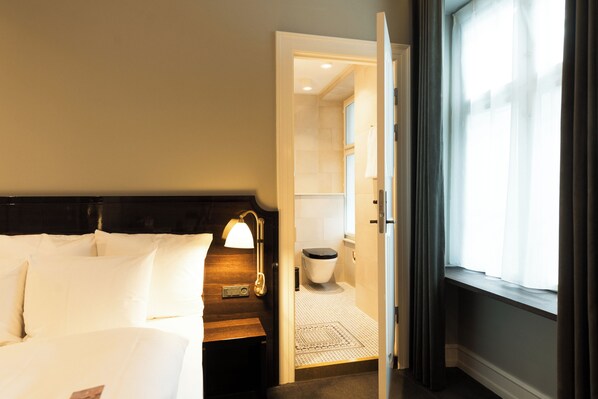 Una habitación de hotel con una cama, una mesita de noche, una lámpara, un ventanal con cortinas y un baño visible a través de una puerta abierta.