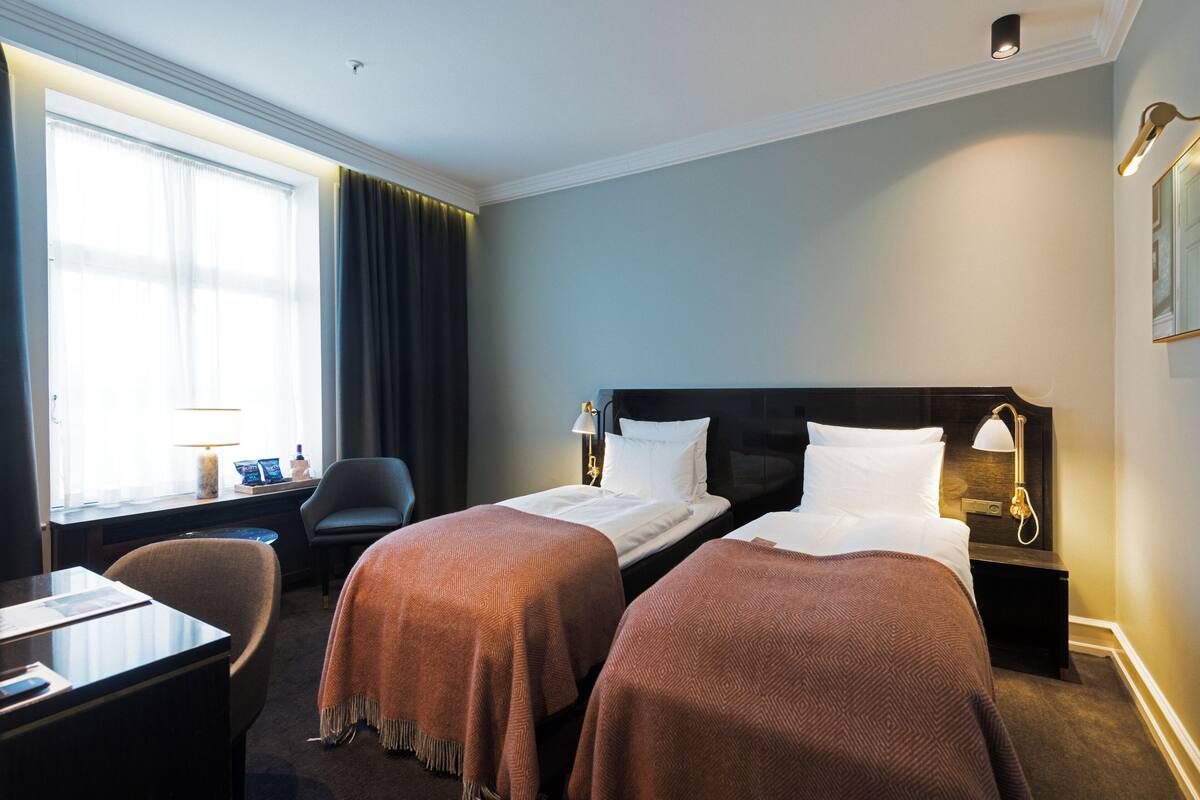 A hotel room with two beds, a desk, a chair, a lamp, and a window with curtains.