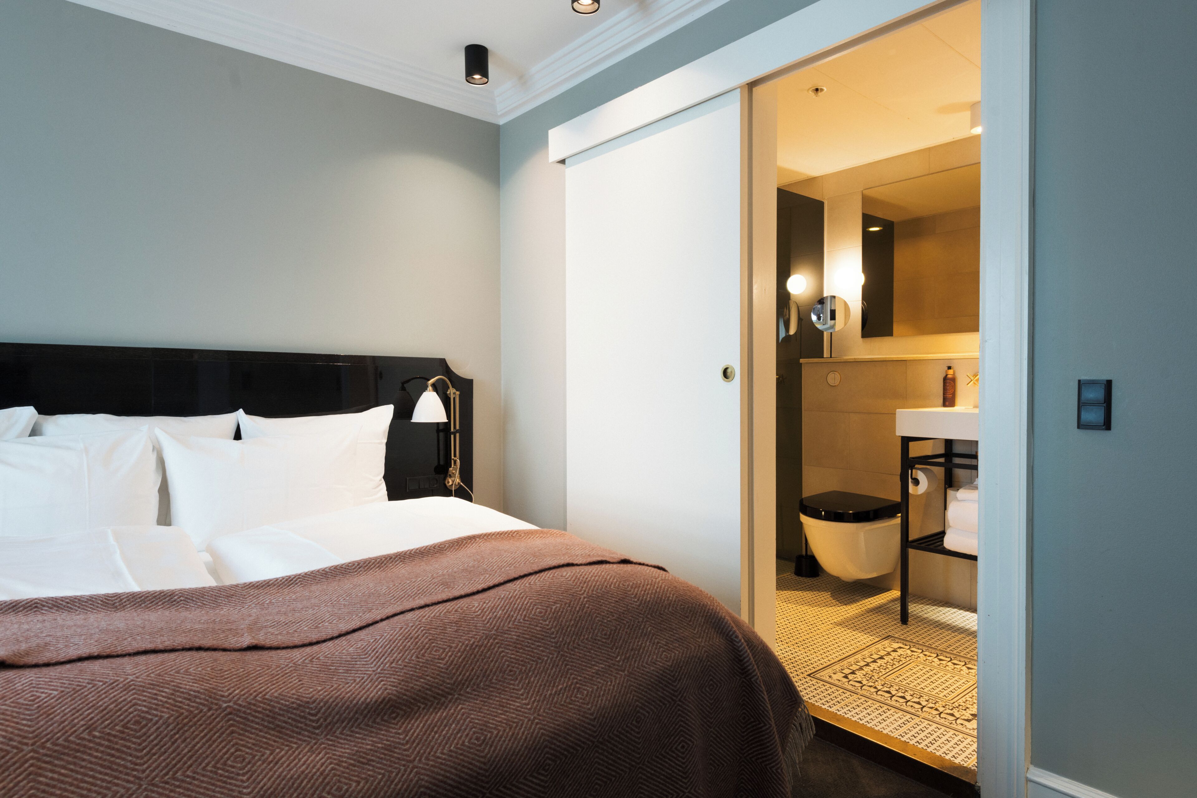 Une chambre d’hôtel comprenant un lit, une table de chevet, une lampe, une salle de bain avec des toilettes, un lavabo et un miroir.