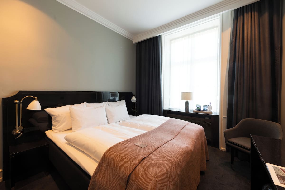 A hotel room with a large bed, a desk, a chair, a nightstand with a lamp, a window with curtains, and a view of the city.