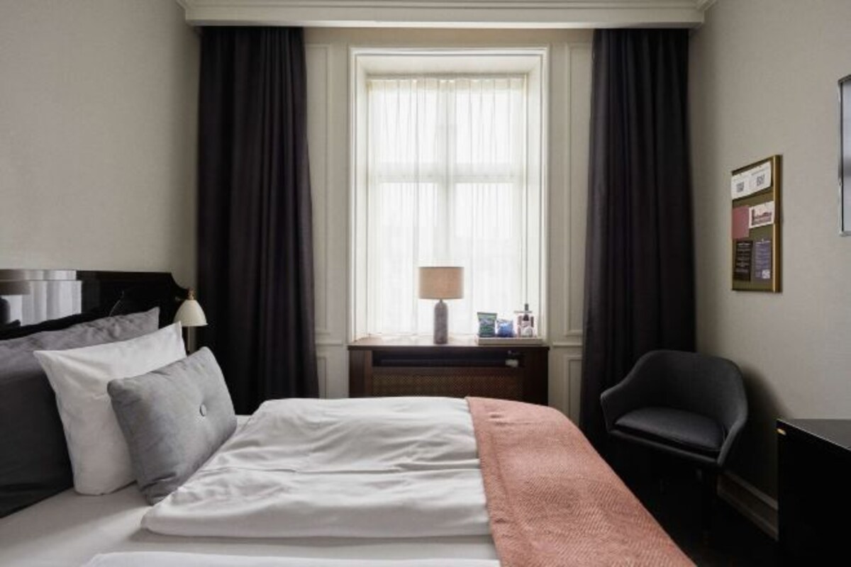 A hotel room with a bed, a desk, a chair, a window with curtains, and a lamp.