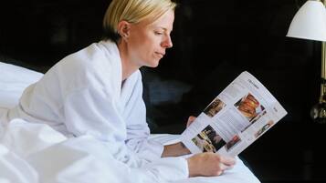 A person sitting on a bed in a room with a dark headboard, reading a magazine.