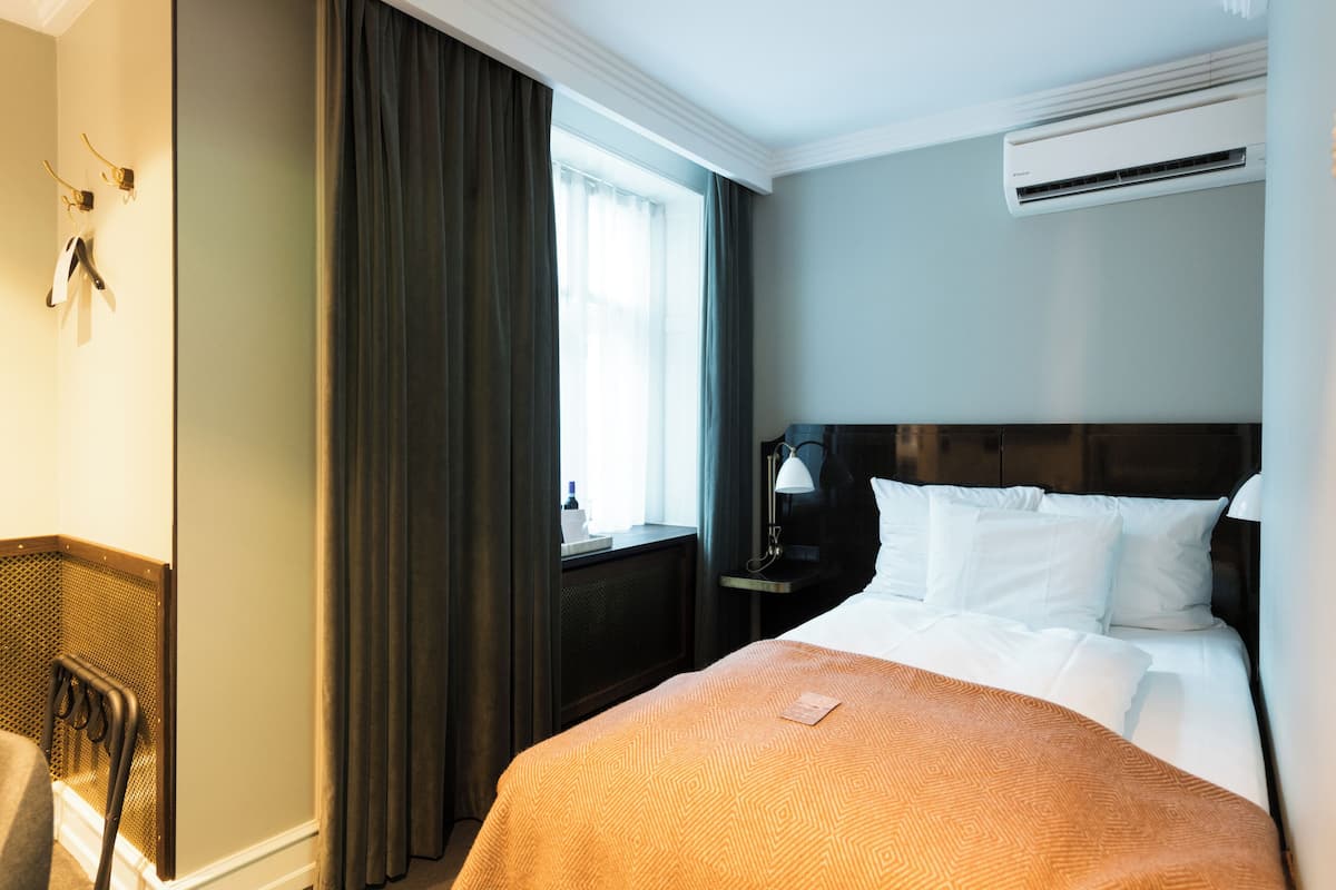A hotel room with a bed, a nightstand, a lamp, a window with curtains, an air conditioning unit, and a chair.