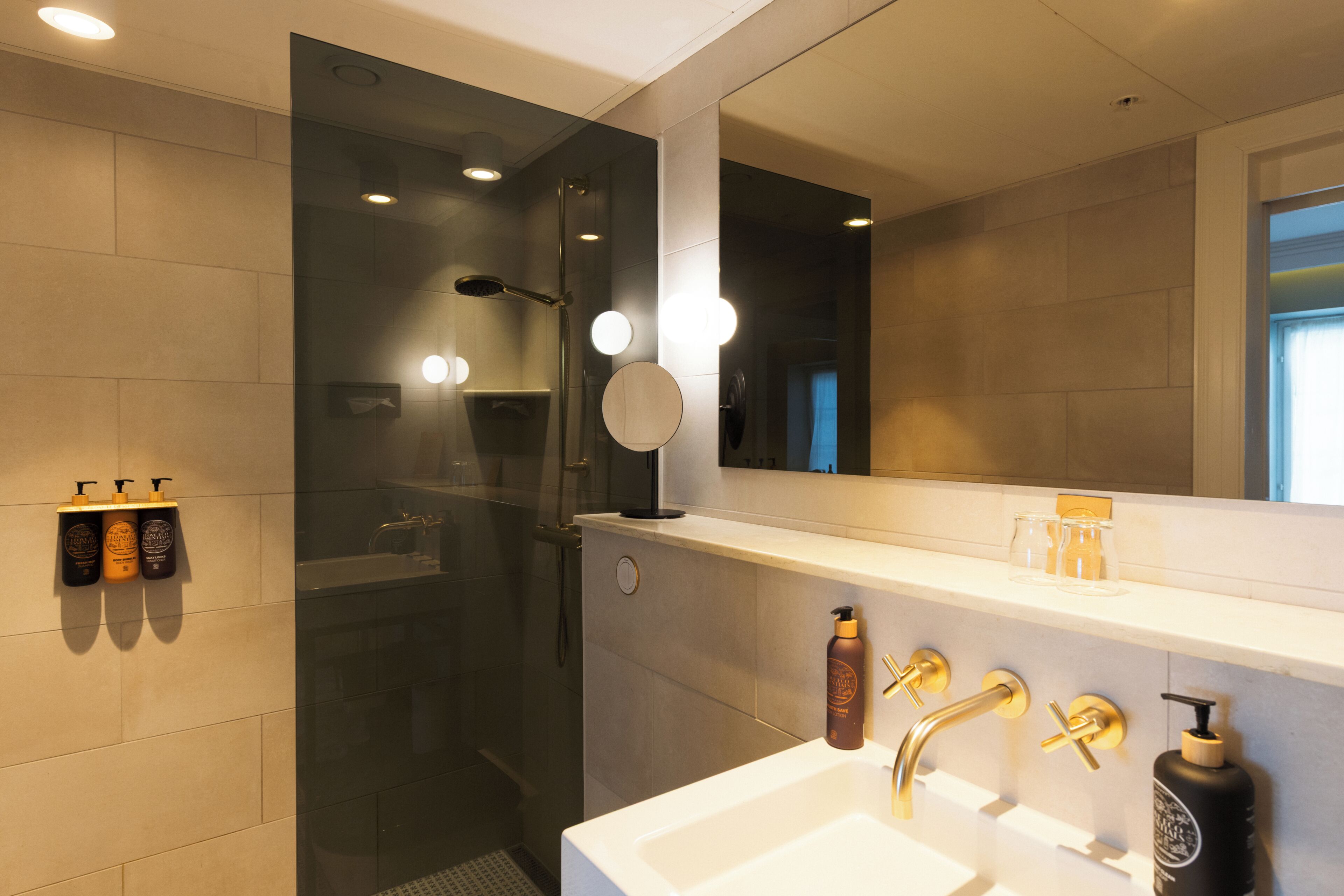 Un baño moderno con cabina de ducha de vidrio, un espejo grande y un lavamanos blanco con detalles dorados.
