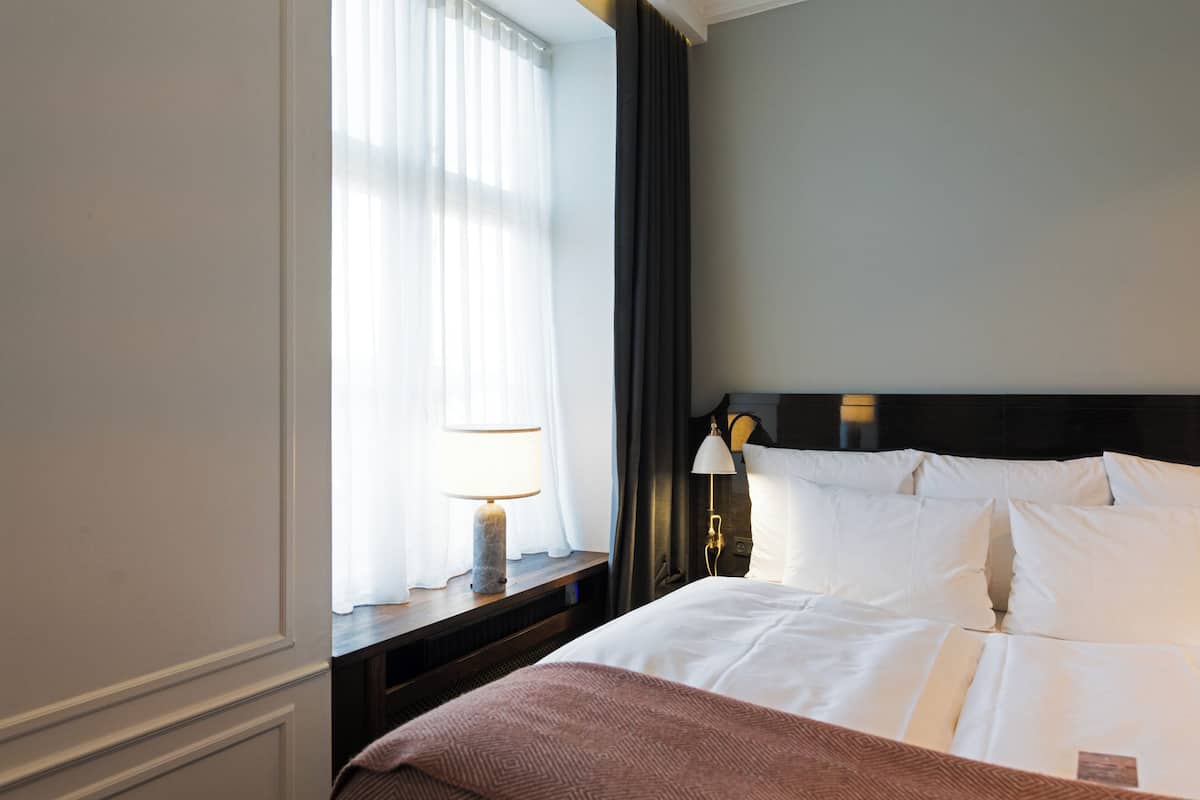 A hotel room with a bed, a nightstand with a lamp, a window with curtains, and a dark headboard.