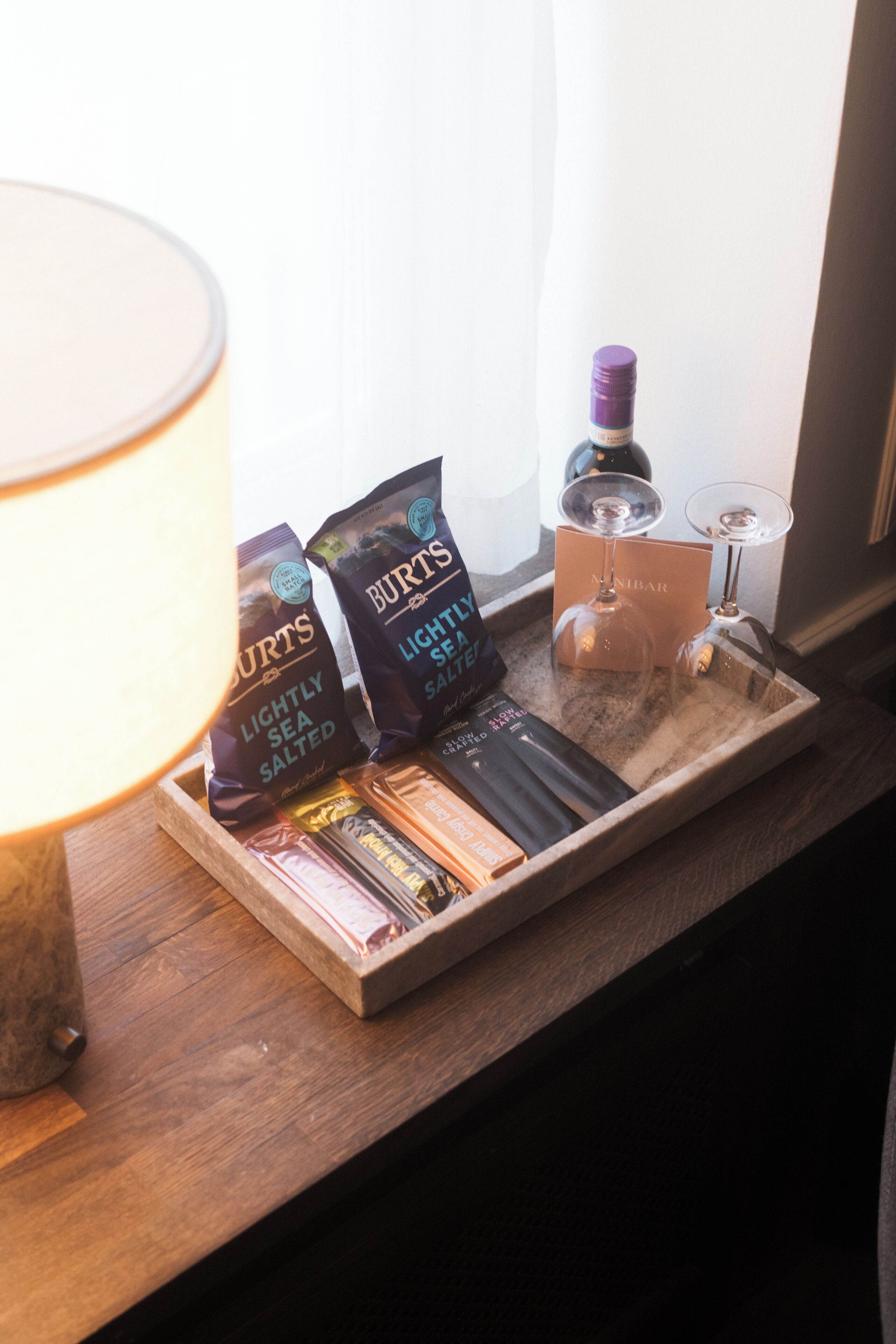 A tray with packaged snacks, a bottle of wine, and two wine glasses on a wooden surface near a window.