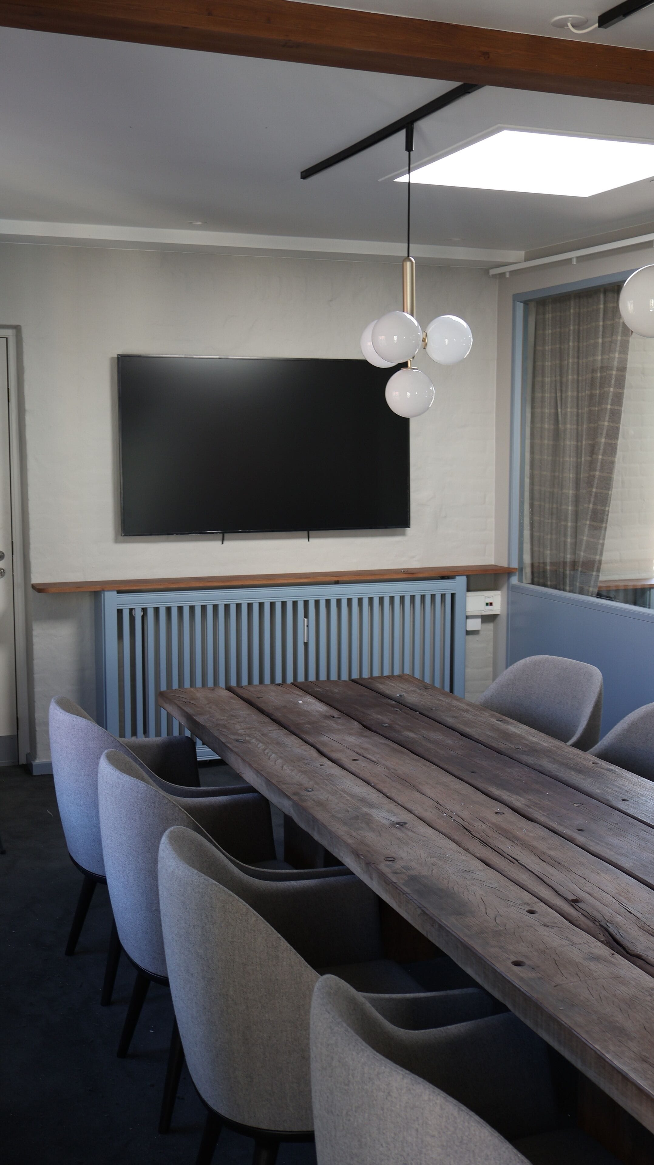 A modern dining area with a long wooden table, grey upholstered chairs, a black flat-screen TV mounted on the wall, and a pendant light fixture.