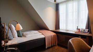Een knus, klein kamertje met een bed, een bureau en een stoel. Er is een raam met gordijnen, een lamp en een klein tafeltje met een fles en snacks.