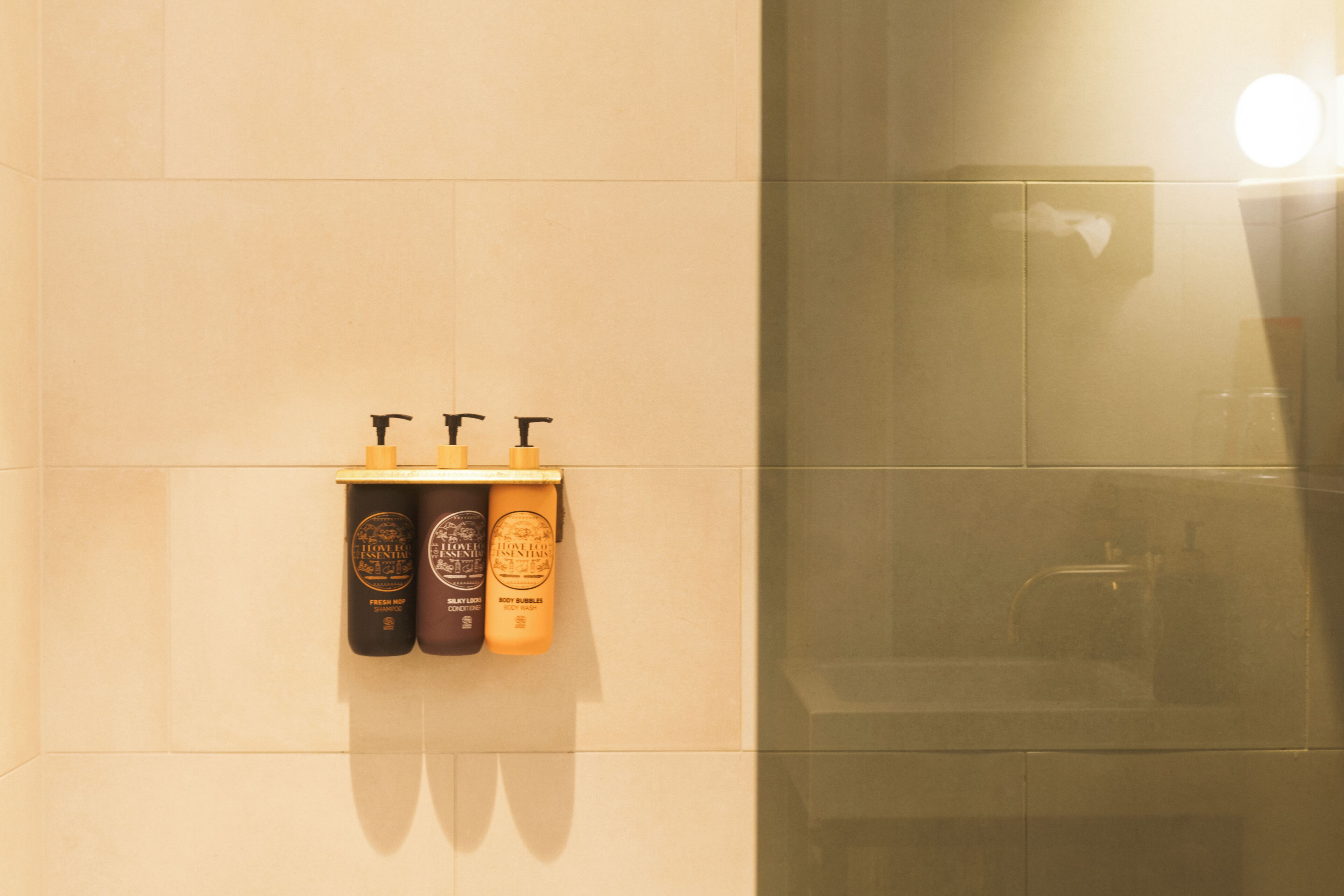 A bathroom with a mirror, a hand dryer, and three bottles of toiletries on a wall-mounted shelf.