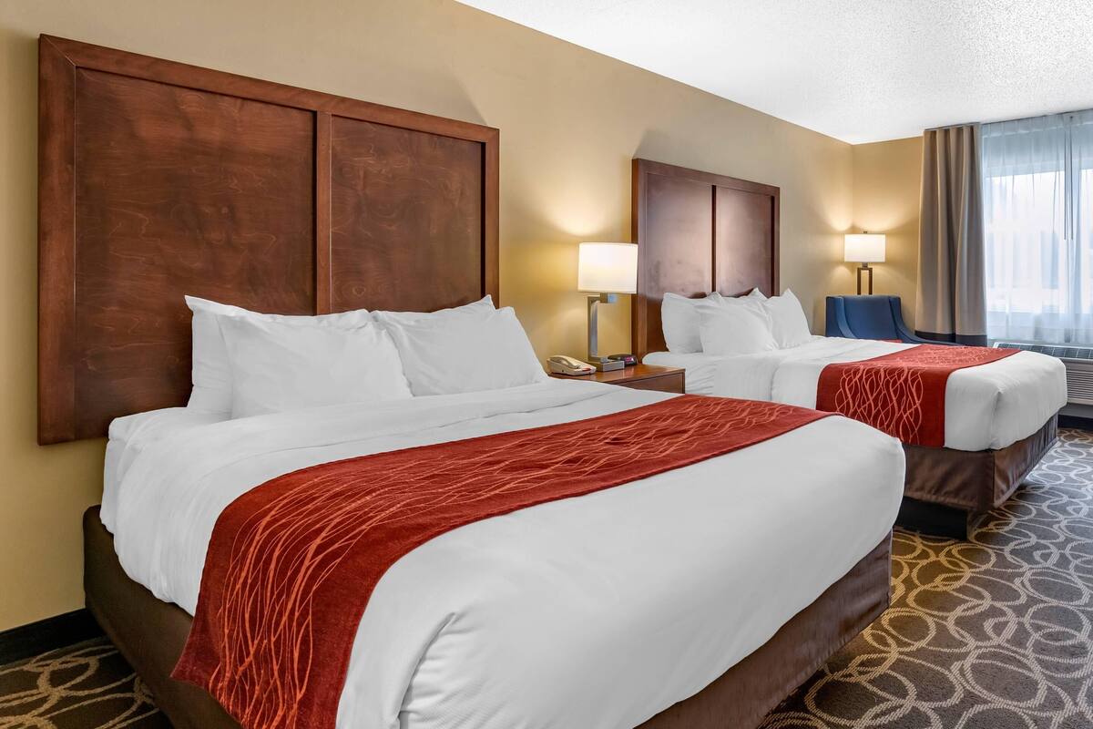 A hotel room with two beds, each with white bedding and a red throw blanket. There are wooden headboards, a nightstand with a lamp, and a large window with curtains.