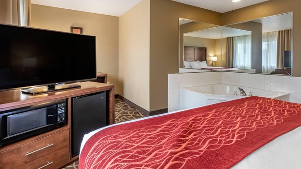A hotel room with a bed, a TV, a minibar, a bathroom with a bathtub, and a large mirror. - Comfort Inn Dyersville Near Field of Dreams (Dyersville)