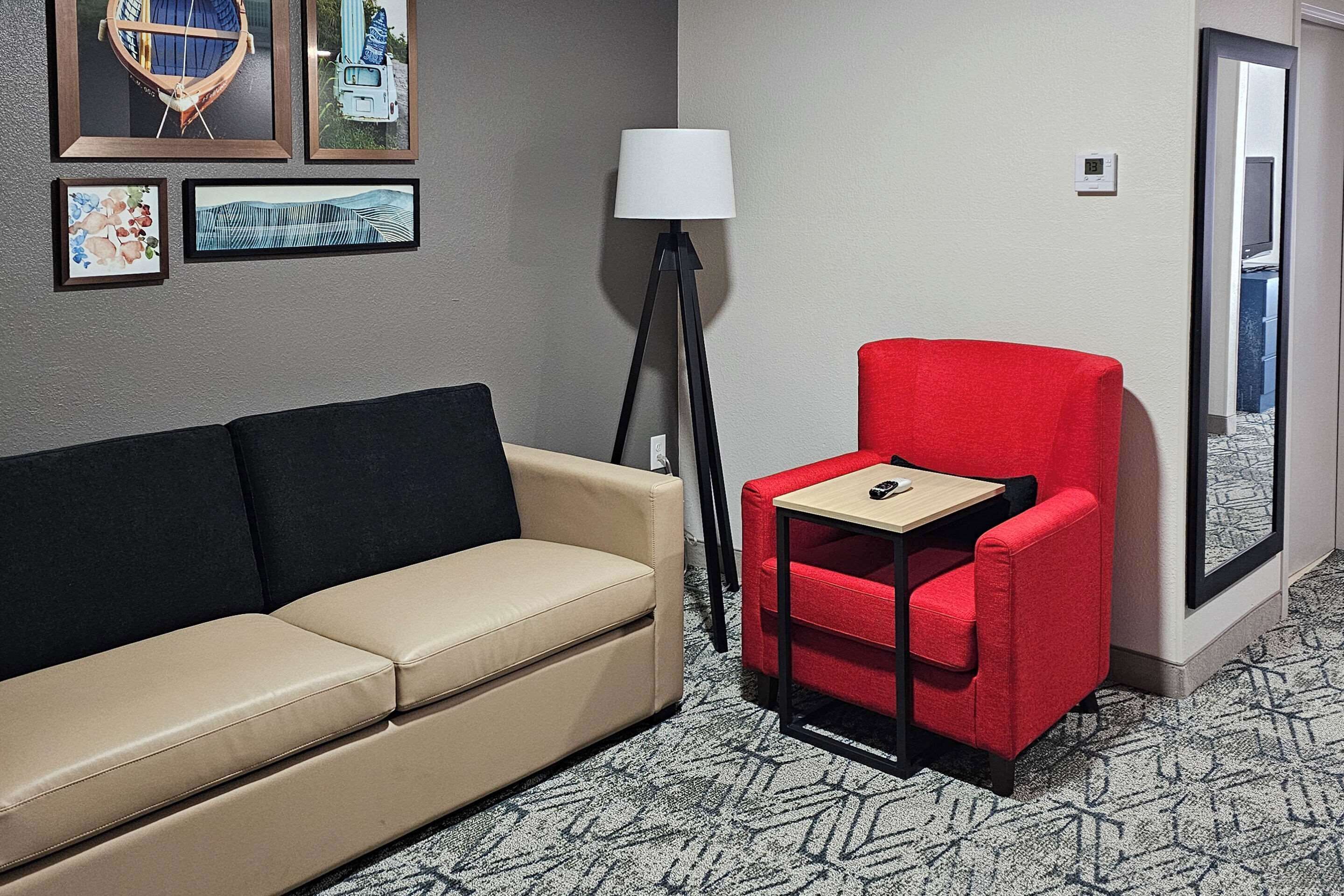 Une chambre d’hôtel avec un canapé beige, un fauteuil rouge, une petite table, un lampadaire et des tableaux encadrés accrochés au mur.