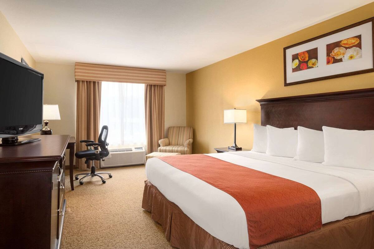 Una habitación de hotel con una cama grande, un escritorio con televisor, una silla, una lámpara y una ventana con cortinas.