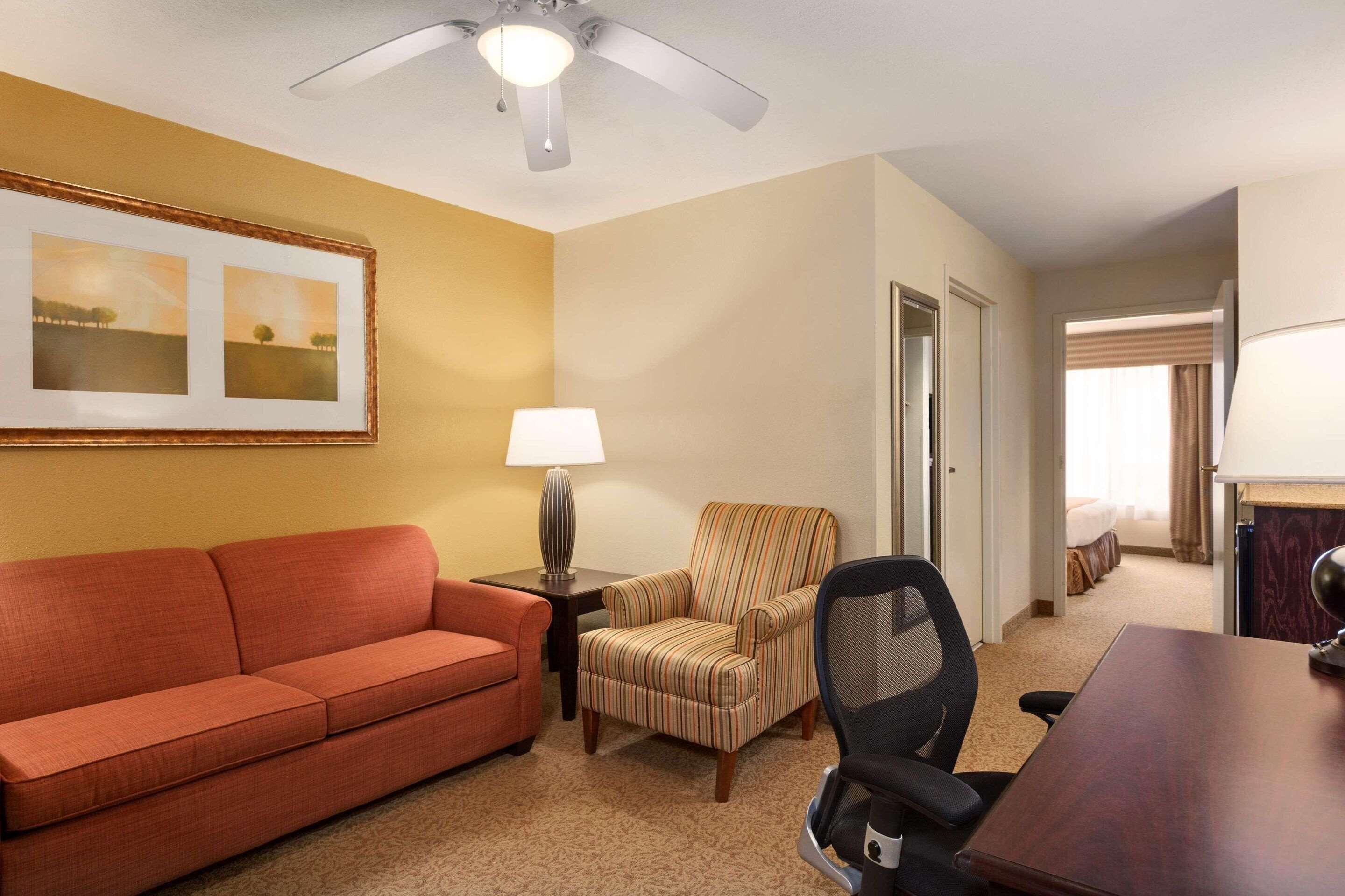 Una habitación de hotel con un sofá rojo, un sillón a rayas, un escritorio con lámpara y un ventilador de techo.
