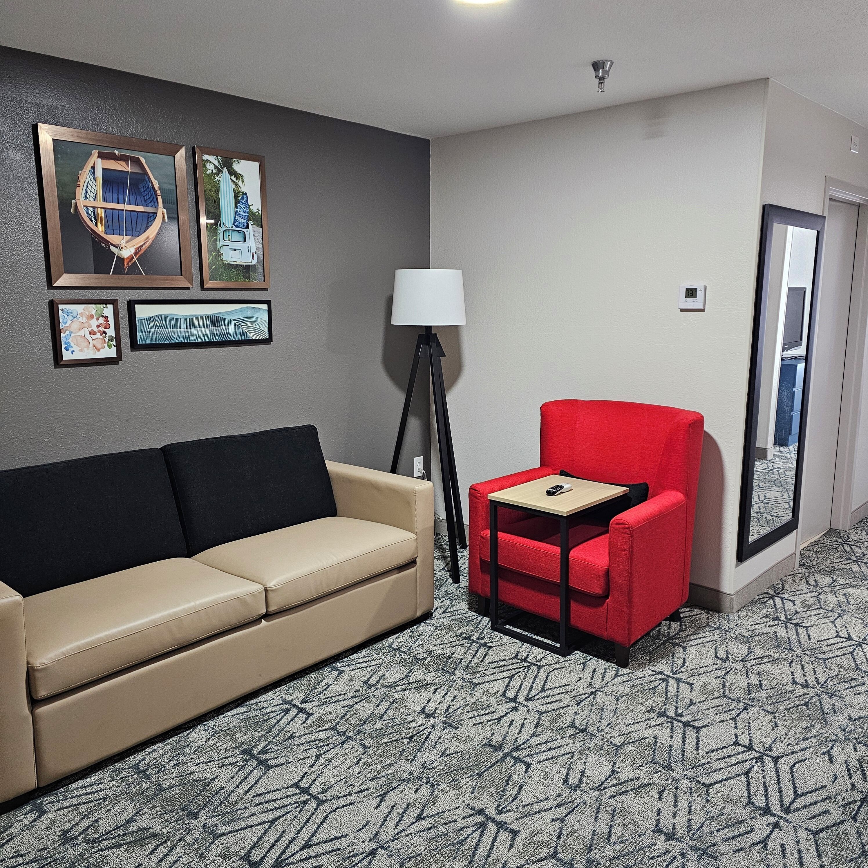 Habitación de hotel con sofá color beige, un sillón rojo, una pequeña mesita de madera, una lámpara de pie y cuadros en la pared.