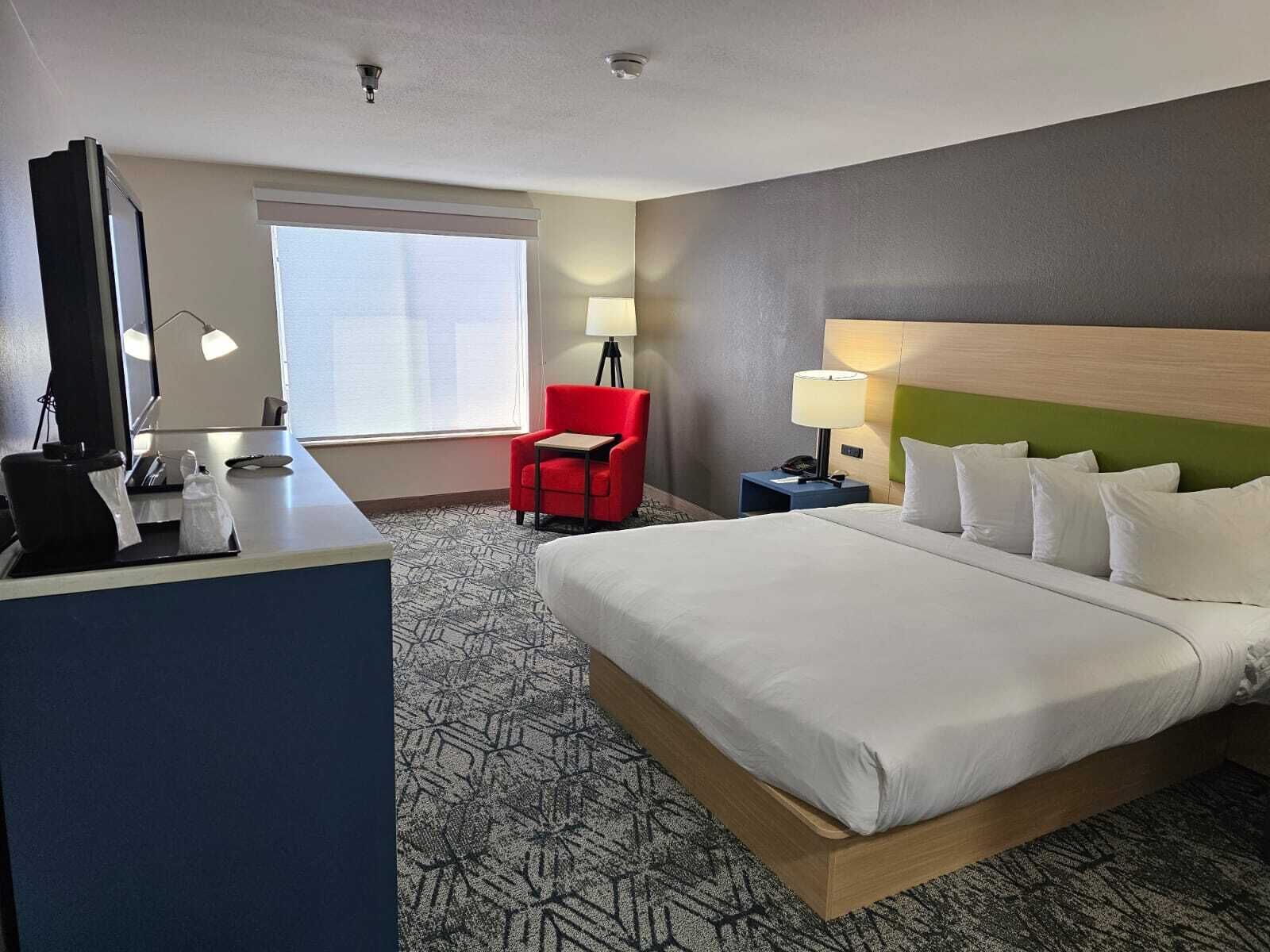 Une chambre d’hôtel avec un grand lit, un bureau avec un ordinateur, une chaise rouge, une table de chevet avec une lampe et une fenêtre avec des stores.