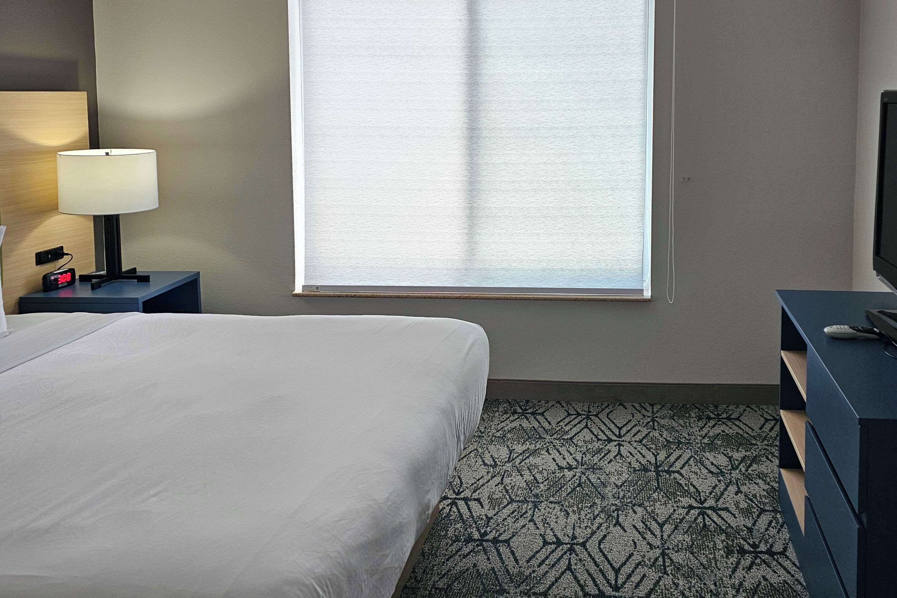 Habitación de hotel con cama, mesita de noche con lámpara, televisión y ventana con persianas.