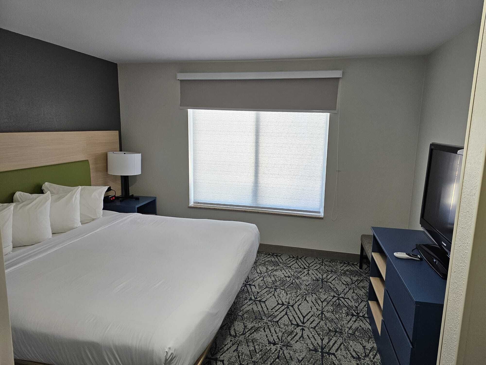 Ein Hotelzimmer mit einem großen Bett, einem Fernseher, einem Schreibtisch und einem Fenster mit Jalousien.