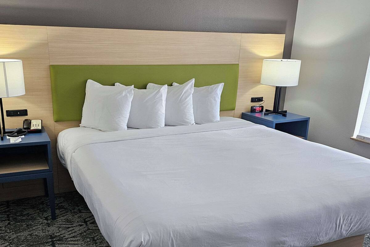 Una habitación de hotel con una cama grande, un cabecero verde, dos mesitas de noche con lámparas y un reloj.