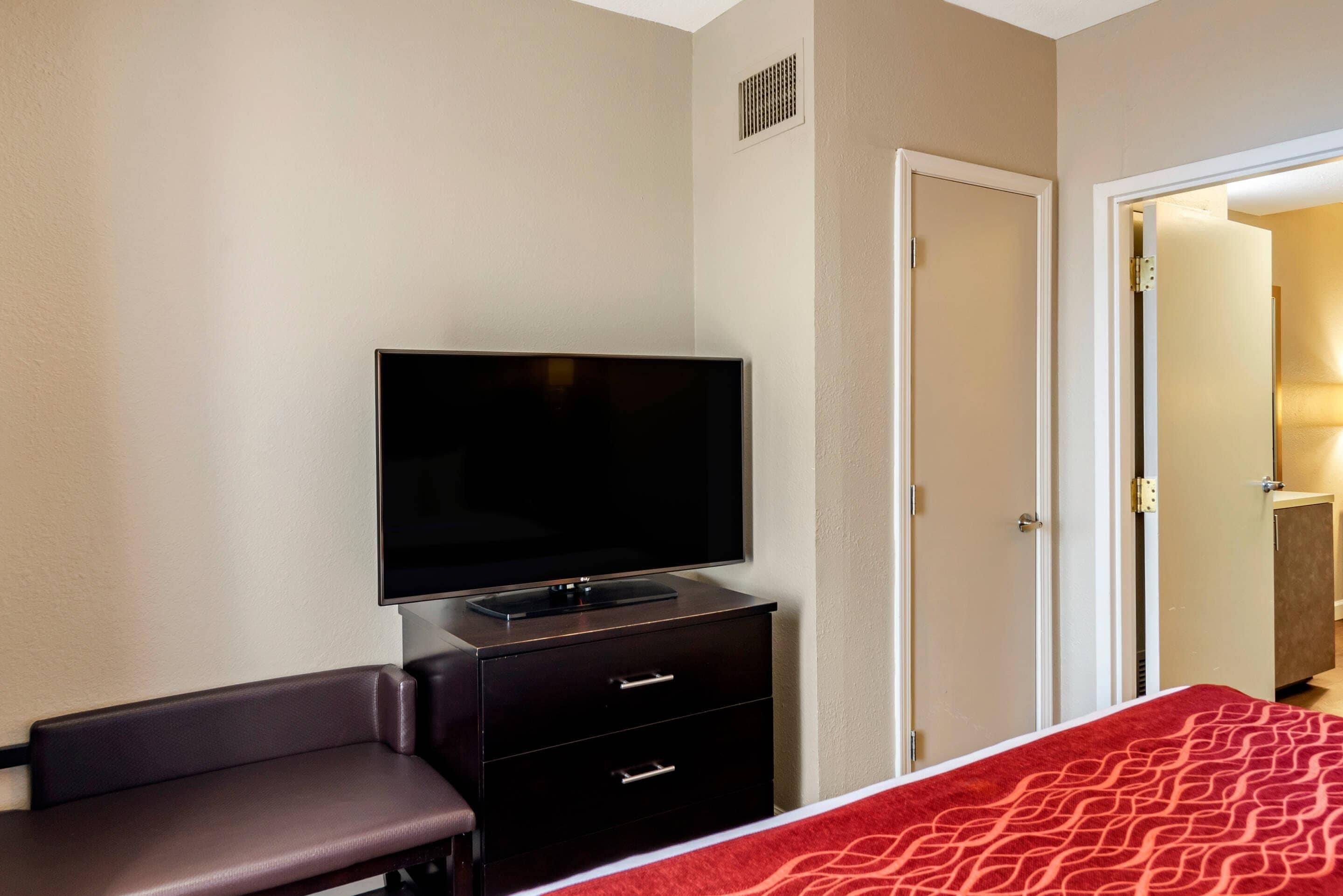 Een hotelkamer met een flatscreen televisie op een dressoir, een rood bed met een patroonspreidek en een deur naar een andere kamer.