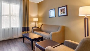 A hotel room with a sofa, a small table, a lamp, and a window with curtains. There are two framed pictures on the wall. - Comfort Inn & Suites near Six Flags (Lithia Springs)