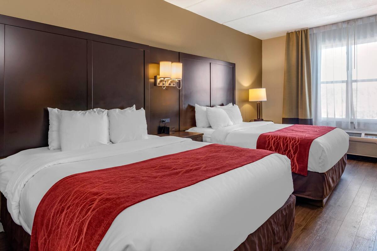 A hotel room with two beds, a large dark wooden headboard, wall-mounted lamps, a window with curtains, and a wooden floor.