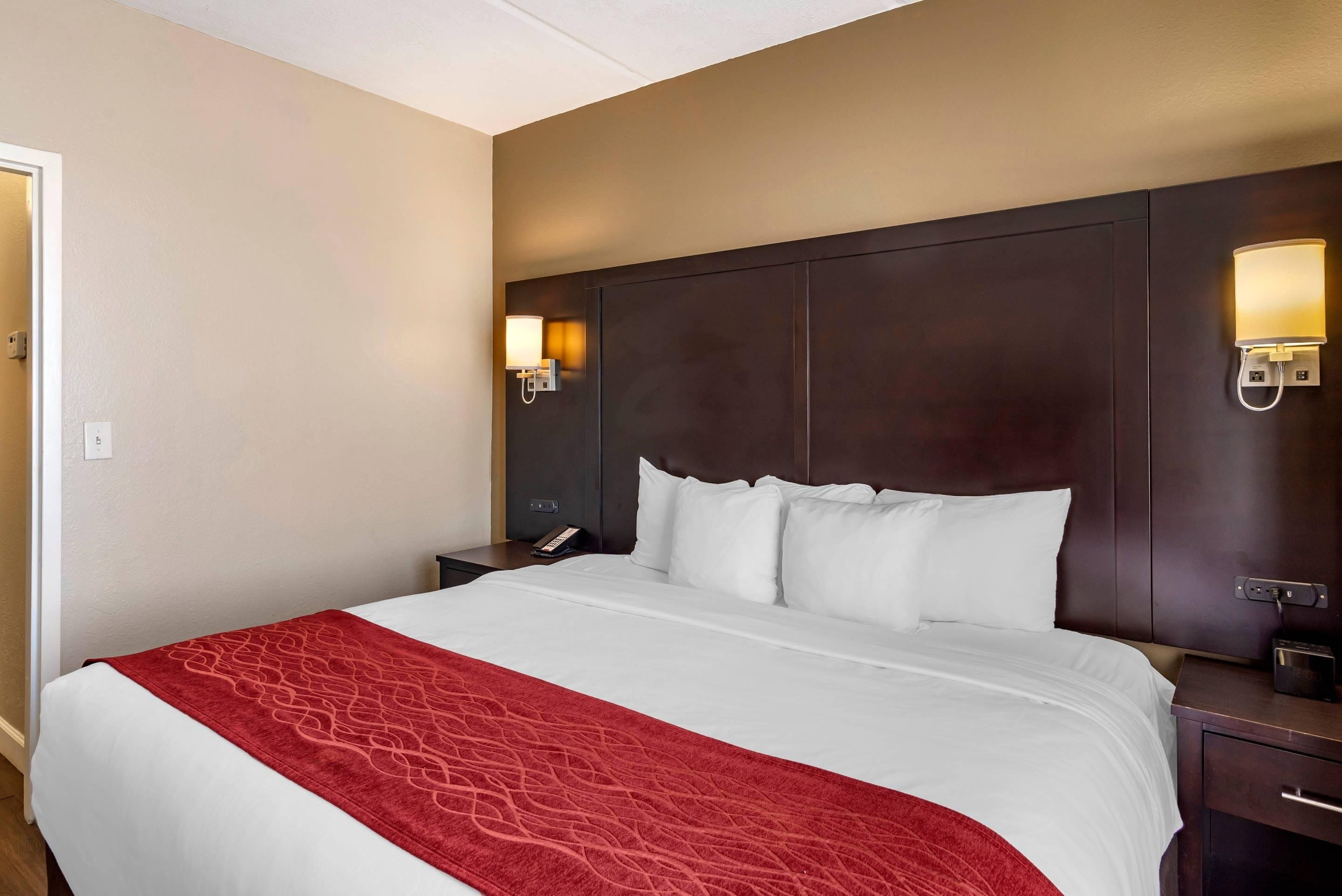 A hotel room with a large bed, a red throw blanket, a dark headboard, and bedside lamps.