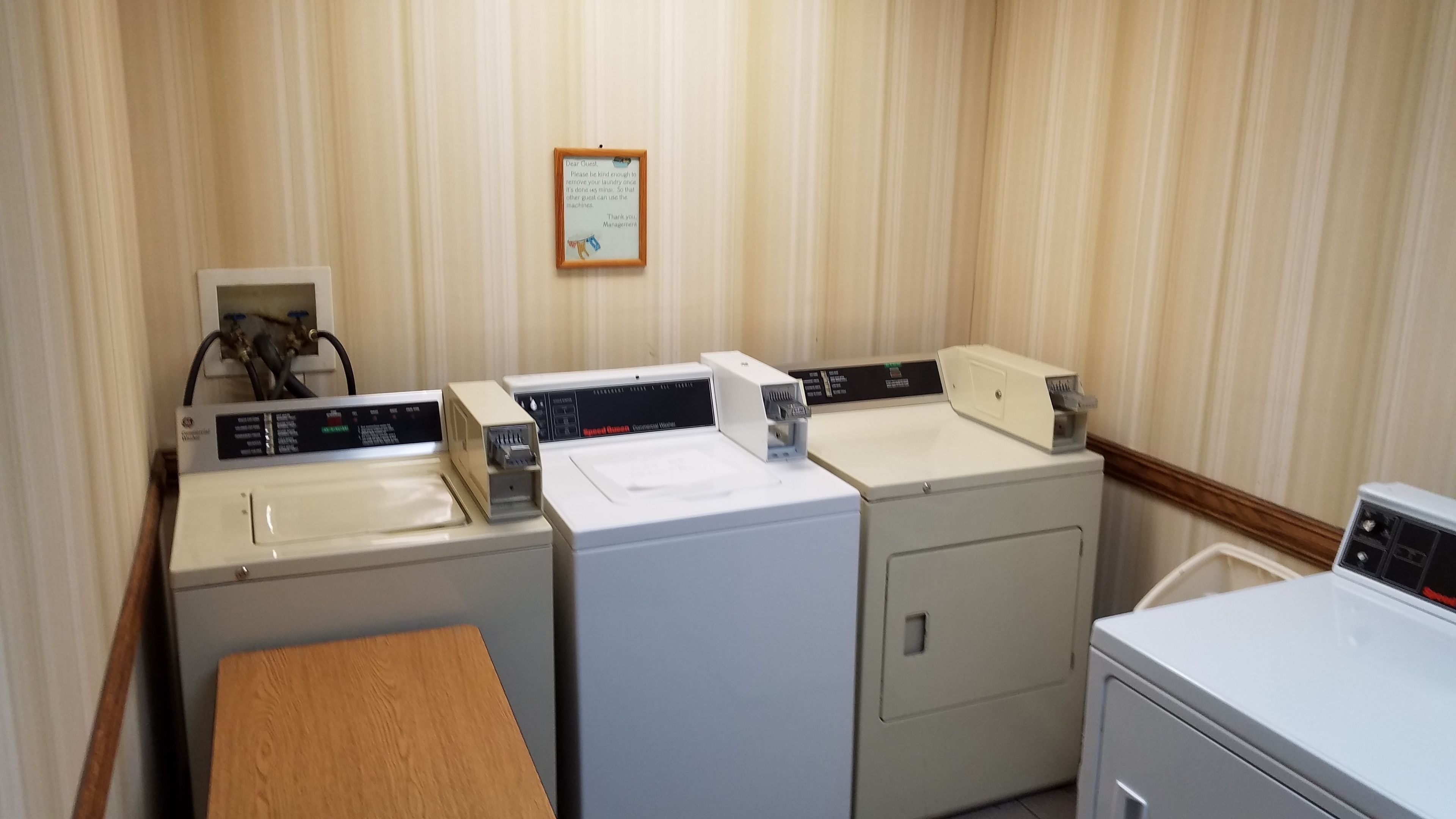 A laundry room with three washing machines and a dryer, a wooden table, and a wall-mounted sign.